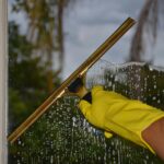Close-up of hand cleaning window with squeegee outdoors using yellow gloves.
